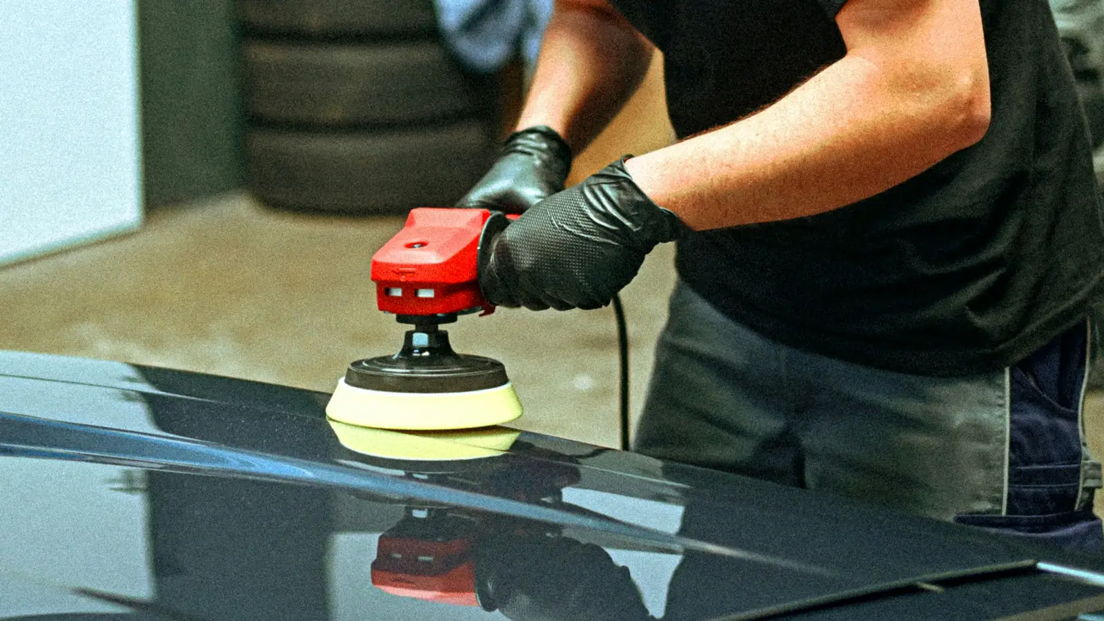 Gloved hands guiding a polisher with a foam pad across a dark car panel